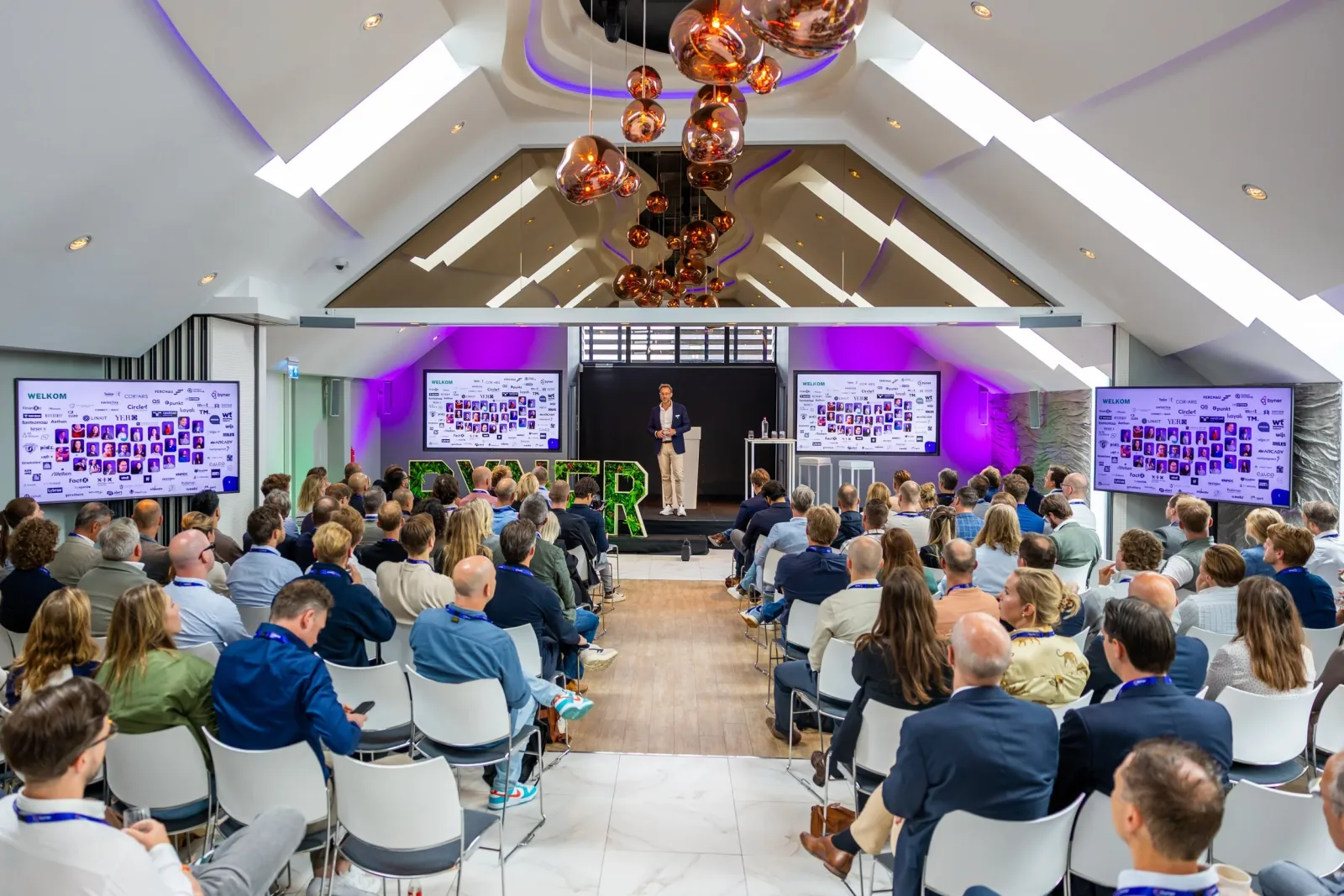 Een grote groep mensen zit in een moderne conferentiezaal, luisterend naar een spreker op het podium. De zaal is licht en ruim, met grote schermen aan beide zijden van de zaal die presentaties weergeven. Er hangen decoratieve, koperen hanglampen aan 