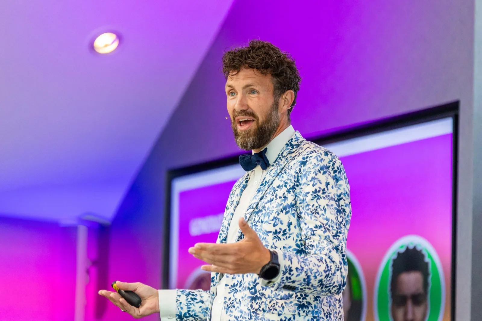 Man met een mannelijke stropdas, in een bloemrijk wit en blauw jasje, die een presentatie geeft in een kamer met roze en blauwe verlichting.